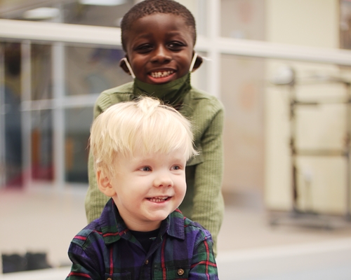 Two little boys smiling one is standing and looking into the camera and the other is sitting and looking away from the camera