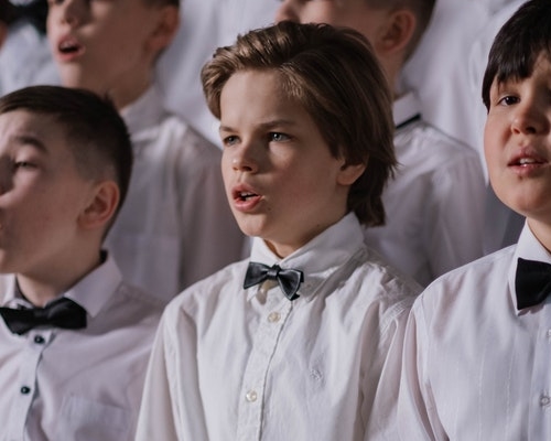 A group of boys wearing white shirts and black bowties with mouths slightly open as if they were speaking or singing