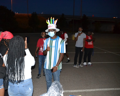 A boy with glasses has a hat on with a fake cake and candles on top and is wearing a mask and has one thumb up while other kids around him are talking