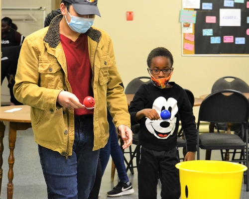 Two boys carrying a spoon with a ball while walking towards a bucket