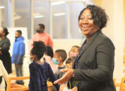 A lady standing behind a group of young children with hand out as a welcoming gesture